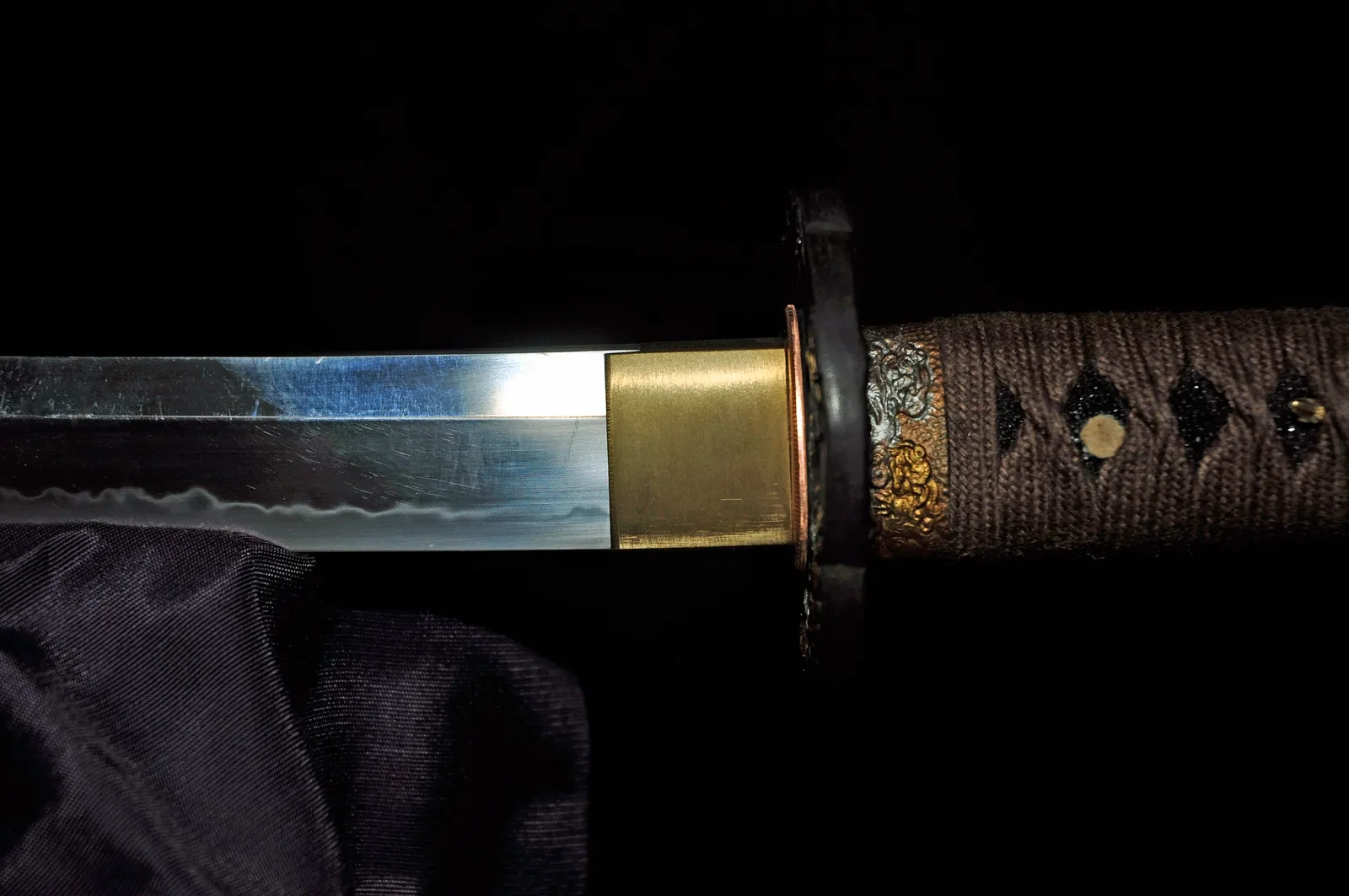 Primer plano de una katana desde el habaki: se aprecia el hamon ondulado a lo largo del filo, la tsuba, la seppa y el arranque del tsuka con su tsuka-ito trenzado.