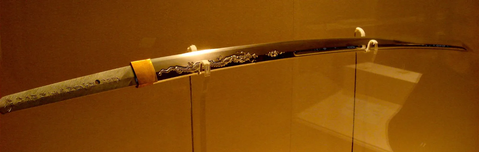 Fotografía de una hoja de katana en vitrina de museo, con un dragón grabado (horimono) junto al habaki, cerca de donde arranca el filo.
