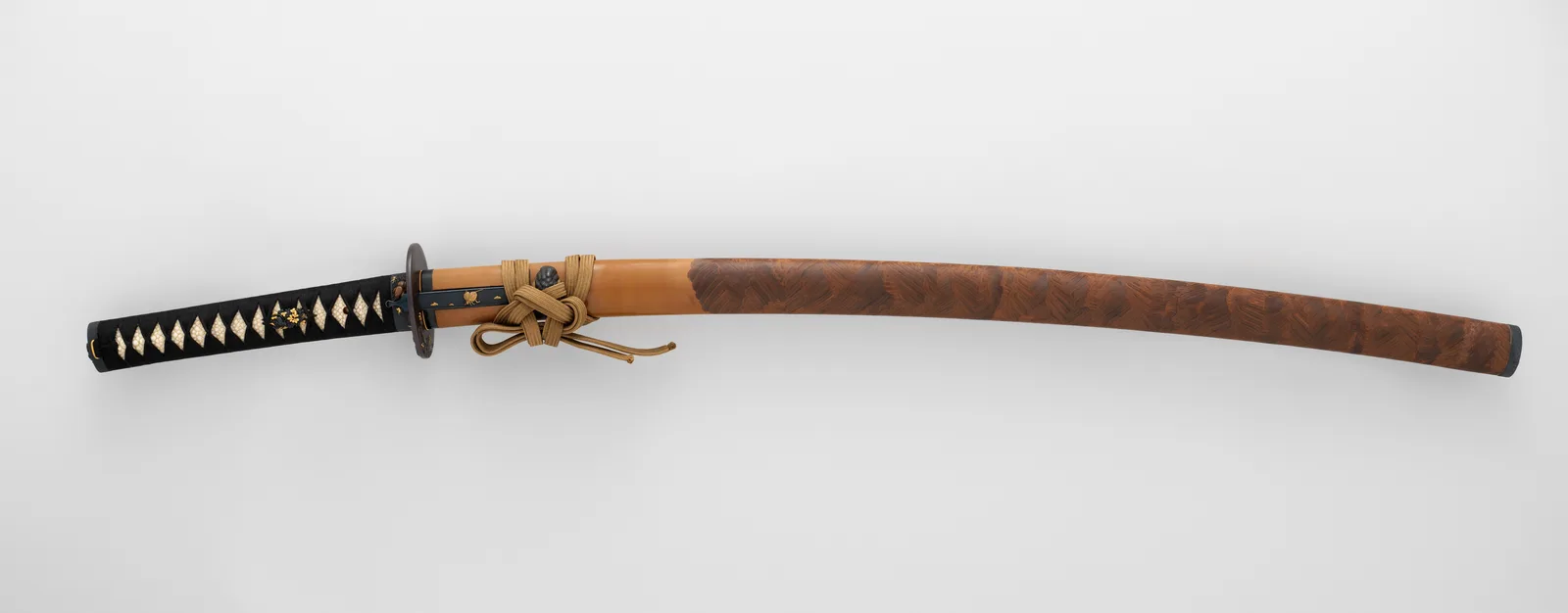 Fotografía de una katana montada en su koshirae completa: empuñadura forrada de piel de raya y trenzado en rombos negro y blanco, tsuba redonda, vaina lacada con textura de veta de madera y cordón sageo anudado junto al kurigata.