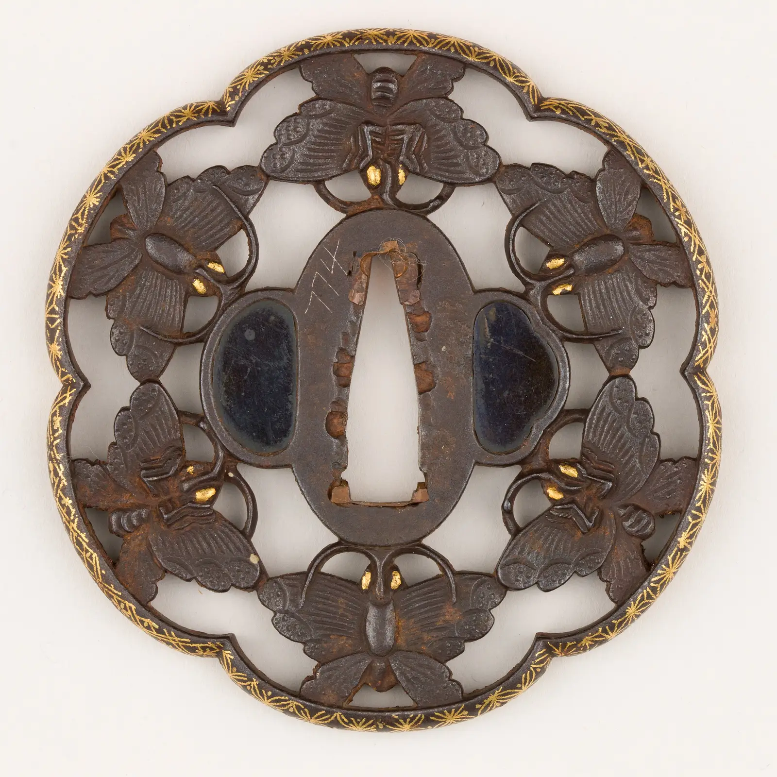 Fotografía de una tsuba de hierro calada (sukashi) con motivo de seis mariposas enlazadas, borde lobulado con incrustaciones de oro y dos óvalos de shakudō a los lados del orificio central para la espiga.