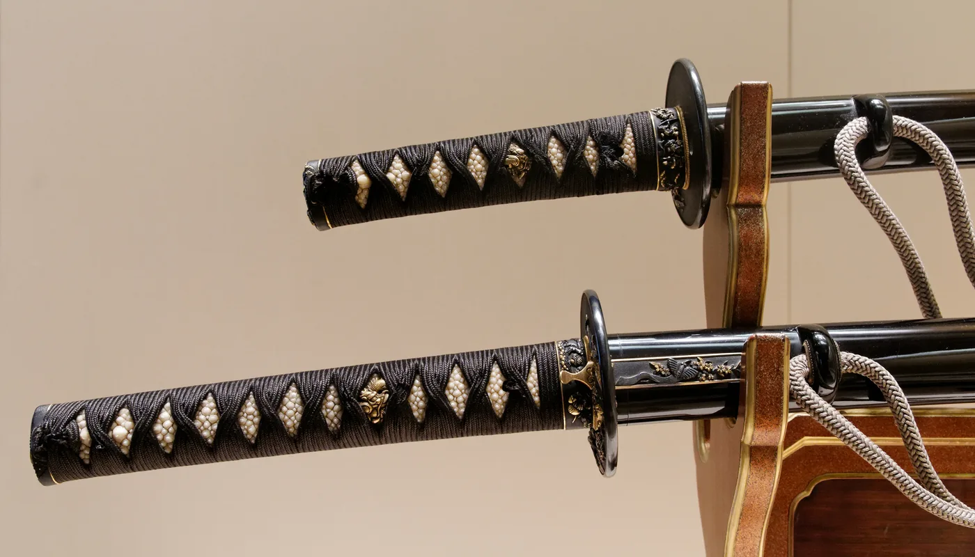 Empuñaduras de un daishō en un soporte, laca negra, piel de raya blanca y trenzado negro con los mismos herrajes en las dos espadas.