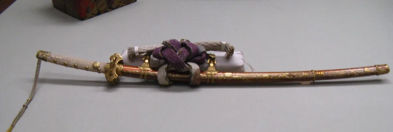 Montura de tachi con vaina lacada en rojo y dorado, cordones de seda blanca y morada anudados junto a la boca de la vaina.