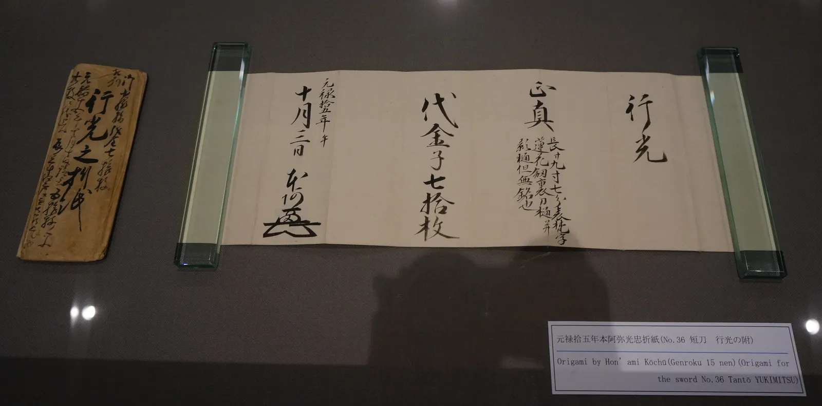 Origami de Hon'ami Kōchū fechado en Genroku 15 (1702), papel doblado con caligrafía vertical que certifica un tantō de Yukimitsu, expuesto en vitrina de museo junto a su funda de madera.