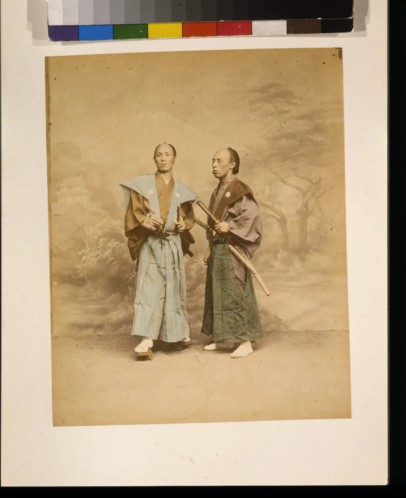 Fotografía de estudio de dos hombres japoneses, de cuerpo entero, cada uno con dos espadas al cinto, coloreada a mano.